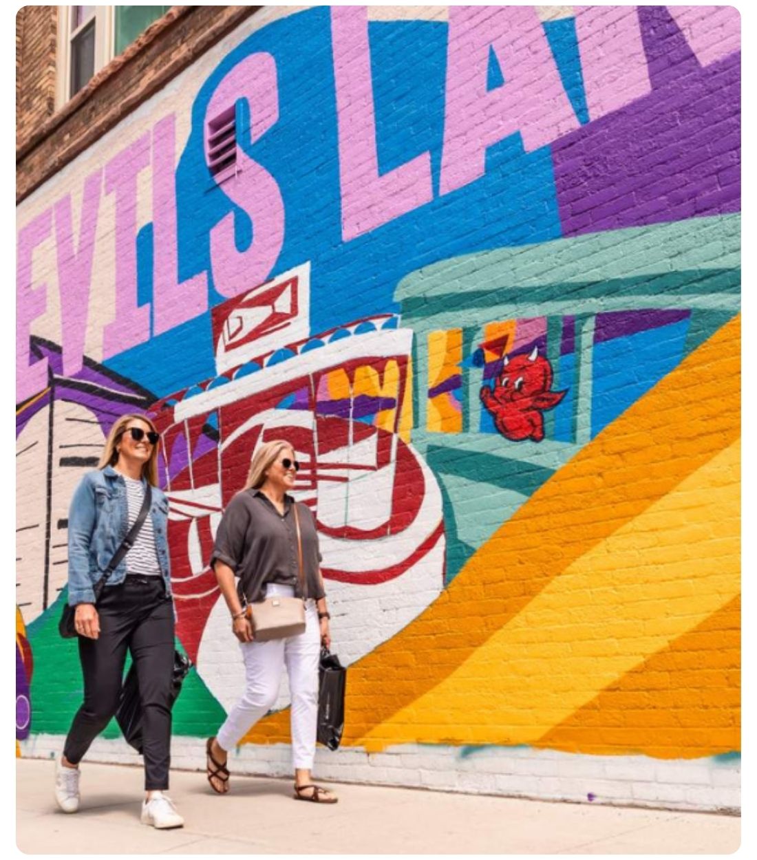 Two people walk past a colorful “Devils Lake” mural painted on a downtown brick wall.