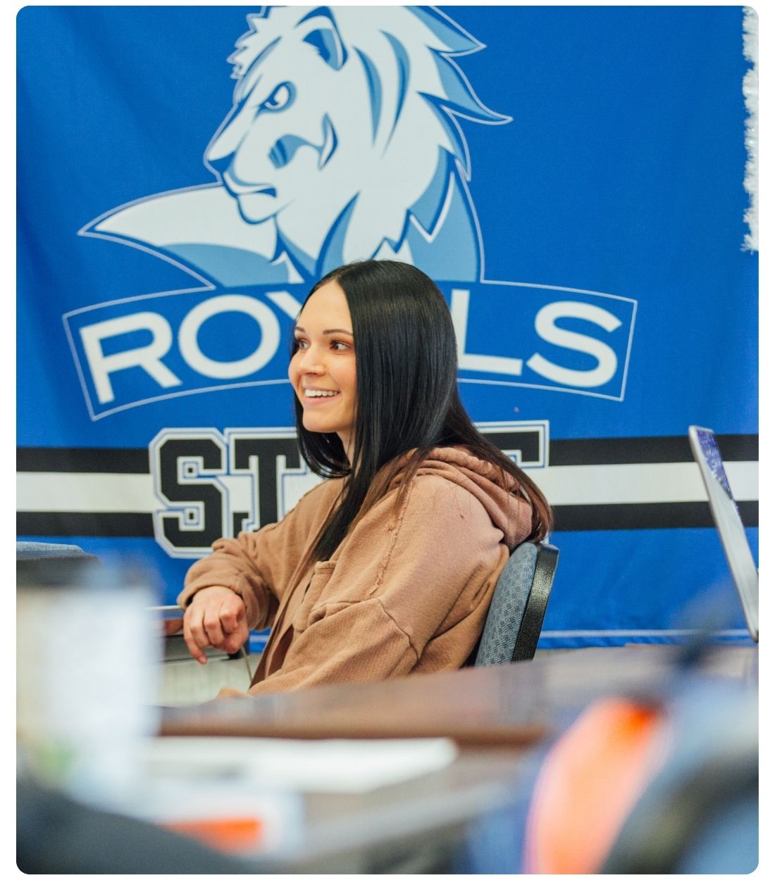 Lake Region State College student sits at a table inside a campus space with a blue Royals State banner in the background.