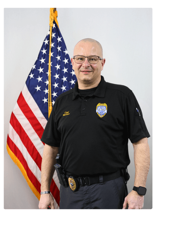 John Maritato standing indoors in front of an American flag, wearing a black polo shirt with a badge patch and duty belt.