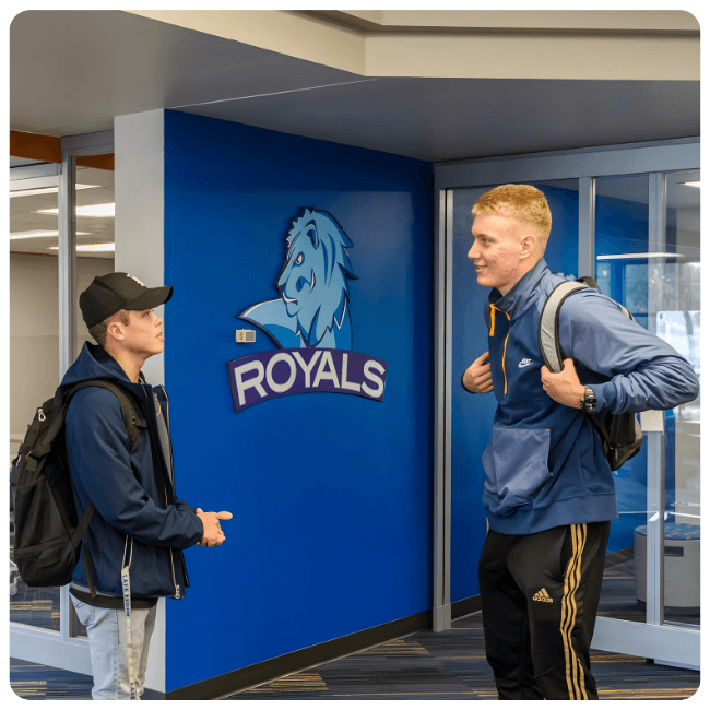 Two Lake Region State College students with backpacks talk inside a campus building near a blue wall displaying the Royals logo.