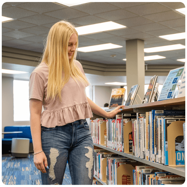Lake Region State College student stands in the campus library browsing books on a shelving unit.