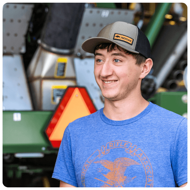 Person wearing a Nodak baseball cap and blue T‑shirt standing in front of farm machinery.