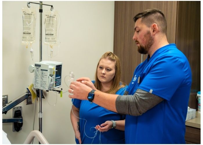 Two students in blue scrubs practice setting up an IV next to an IV pole and medical equipment.