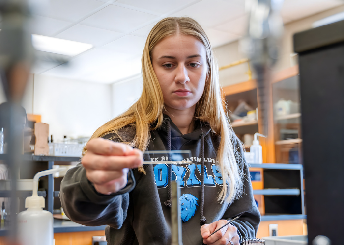 Student uses a dropper to carefully add liquid to a test tube in a science lab, with workstations and equipment visible in the background.