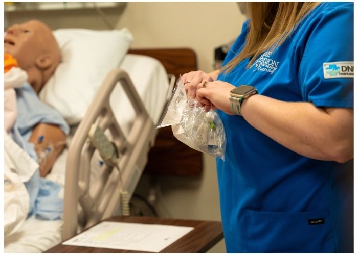 Student in blue scrubs prepares medical supplies next to a hospital bed with a training manikin.