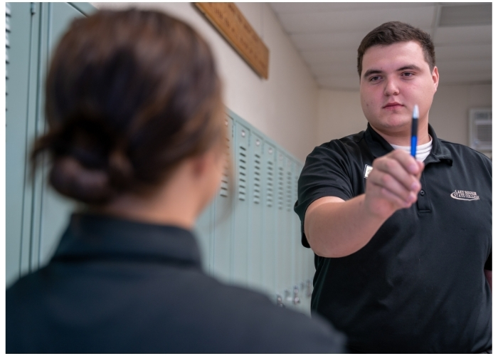 A student extends a pen to another person in a hallway lined with lockers.