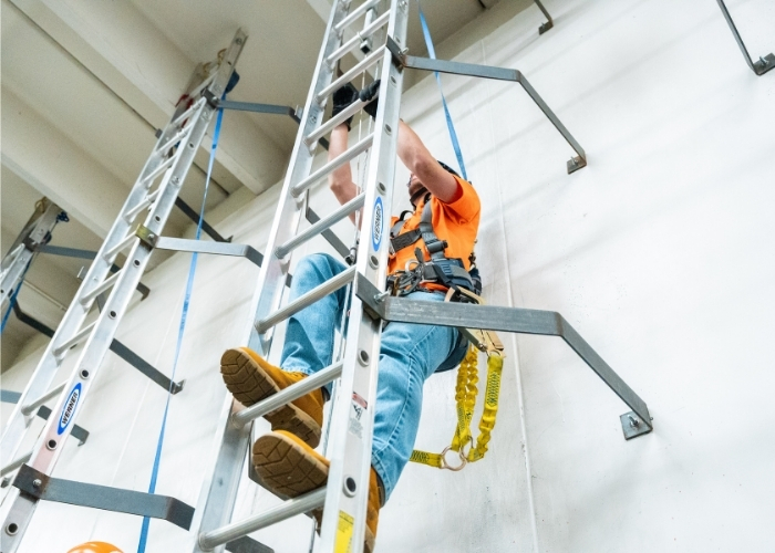 Student wearing a safety harness climbing a training ladder indoors, secured with fall protection equipment for wind energy technician training. Provide your feedback on BizChat