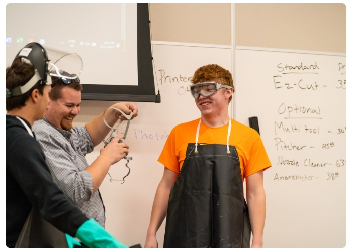 Students wearing safety goggles and protective gear participate in a hands-on lab activity in a classroom.