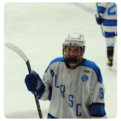 Ice hockey player wearing a black and blue Lake Region State College jersey holding a hockey stick on the ice during a game, with rink boards and spectators visible in the background.