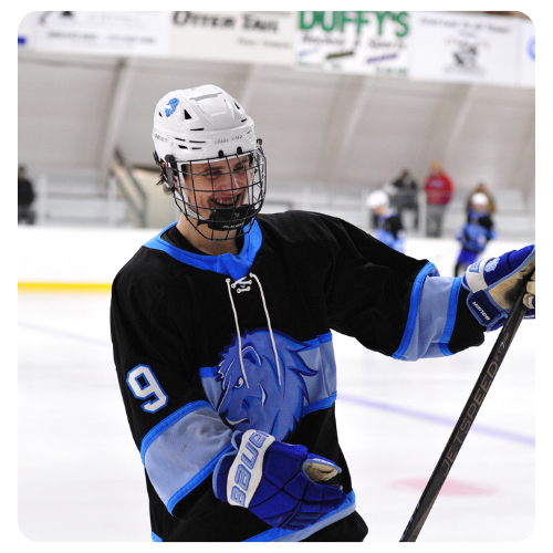 Ice hockey player wearing a black and blue Lake Region State College jersey with the number 9, holding a hockey stick on the ice during a game, with rink boards and spectators visible in the background.