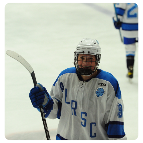 Ice hockey player wearing a black and blue Lake Region State College jersey holding a hockey stick on the ice during a game, with rink boards and spectators visible in the background.