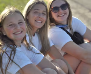 Three people sitting outdoors on the ground wearing white shirts, facing the camera.