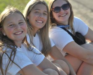 Three people sitting outdoors on the ground wearing white shirts, facing the camera.