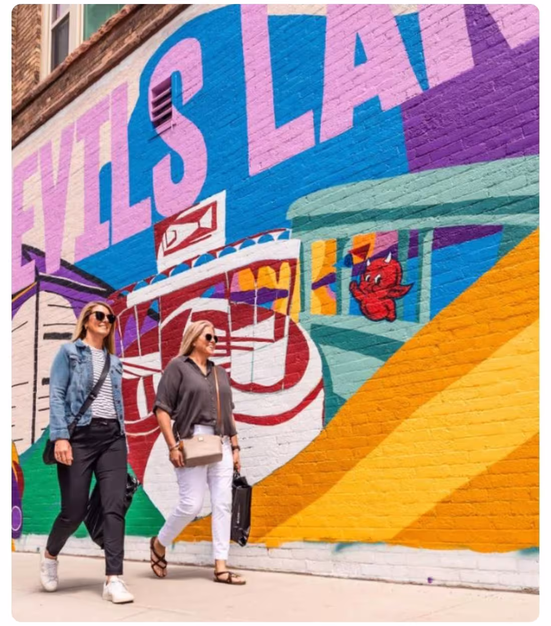 Two people walk past a colorful “Devils Lake” mural painted on a downtown brick wall.