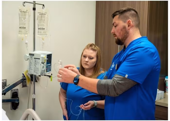 Two students in blue scrubs practice setting up an IV next to an IV pole and medical equipment.