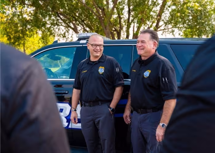 Two law enforcement instructors stand beside a patrol vehicle during an outdoor training session.