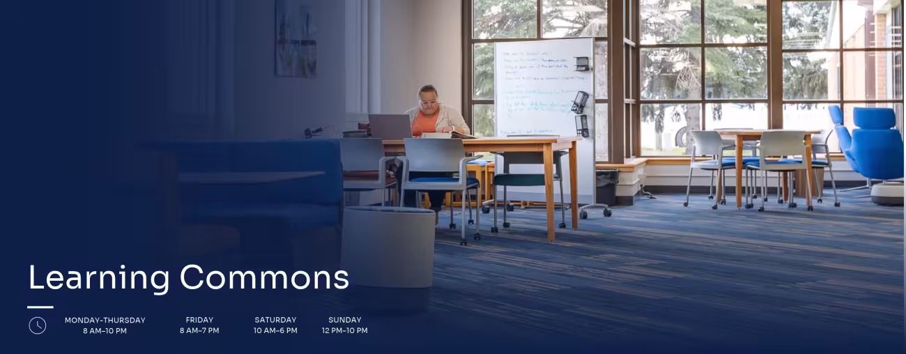 Student studying at a table in the Learning Commons, a collaborative campus space with study tables, seating areas, and large windows.