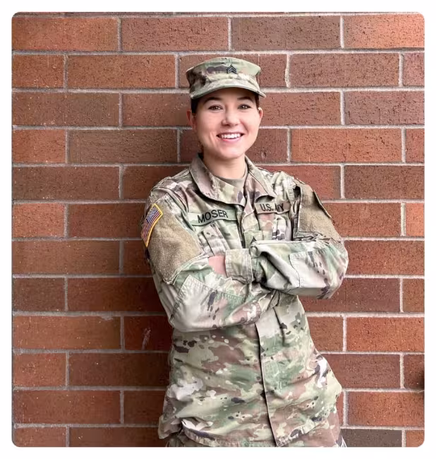 Person in military uniform standing with arms crossed in front of a brick wall.