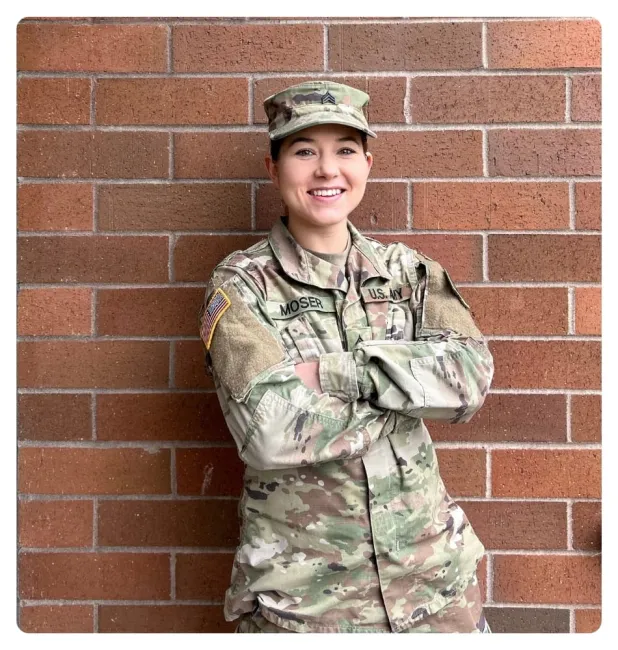 Person in military uniform standing with arms crossed in front of a brick wall.