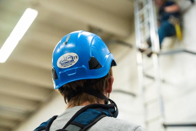 A wind energy student in safety gear watching classmate climb